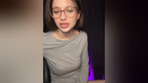 Media: Video of a fair-skinned woman with light brown hair and glasses, wearing a gray long-sleeve top, sitting in a dimly lit room with purple lighting.