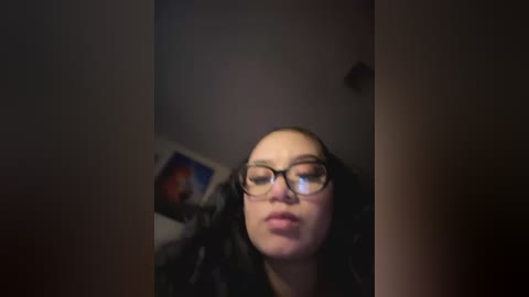 Media: Video of a young woman with light skin and straight black hair, wearing glasses, making a pouty face. Background is dimly lit with a blurry, indistinct wall and a framed picture hanging on it.