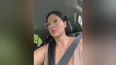 Media: A video of a woman with long black hair, wearing glasses and a beige top, driving in a car with a seatbelt, blurred greenery visible outside.