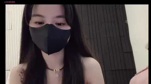 Media: A video of an East Asian woman with long black hair, wearing a black face mask and a simple necklace, standing in front of a dark wooden door.