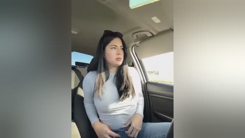 Media: Video of a young woman with long, straight black hair and a light complexion, wearing a white long-sleeved top and blue jeans, sitting in the passenger seat of a car, looking contemplative.