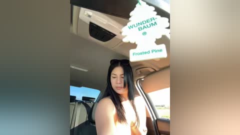 Media: Video of a woman with long black hair, wearing sunglasses and a topless, smiling, seated in a car with air fresheners labeled \"Wunderbaum\" and \"Frosty Pine\" hanging from the rearview mirror.