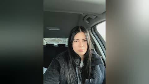 Media: Video of a young woman with long black hair, wearing a dark jacket, seated in a car with a neutral gray interior.