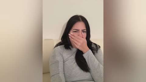 Media: A video shows a woman with long black hair, wearing a gray sweater, covering her mouth with her hand, sitting on a beige chair against a plain, light-colored wall.