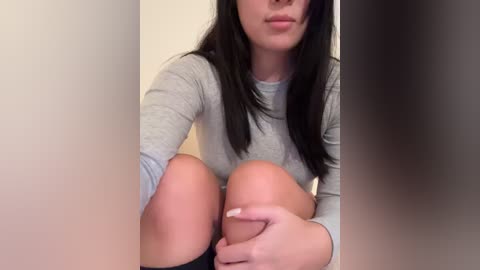 Media: Video of a young woman with fair skin, long black hair, and light gray sweater. She is sitting with her knees pulled up to her chest, holding her right knee with her left hand. Background is a plain, off-white wall.