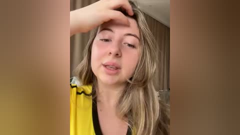 Video of a young Caucasian woman with light skin and long blonde hair, wearing a yellow top, wiping her forehead with her hand. Background shows blurred beige curtains.