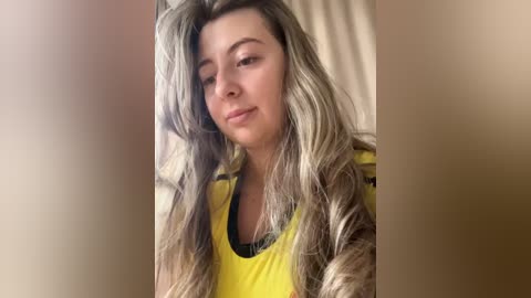 Media: Video of a fair-skinned woman with long, wavy blonde hair, wearing a yellow tank top. She has a neutral expression, with a soft, natural lighting background.