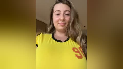 Media: A video of a young woman with long, wavy blonde hair, fair skin, and a slightly smiling face. She wears a bright yellow soccer jersey with red accents and black sleeves.