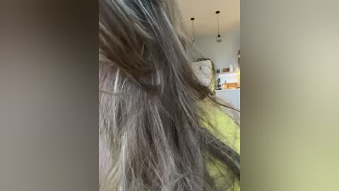 Media: A close-up video of a person's long, messy, dark brown hair blowing in the wind, partially obscuring the face, set against a blurred background of a modern living room with white walls, hanging lights, and a beige couch.