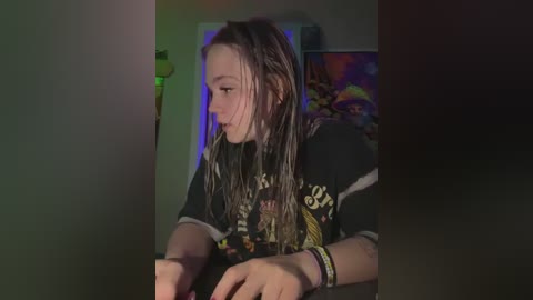 Media: A video of a young, wet-haired woman with fair skin, wearing a dark graphic tee, sitting at a desk in a dimly lit room with colorful, trippy wall art.