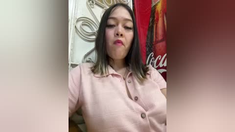 Media: A video of a young Asian woman with shoulder-length black hair, wearing a light pink button-up shirt, puckering her lips. Background features decorative wrought iron and a Coca-Cola poster.