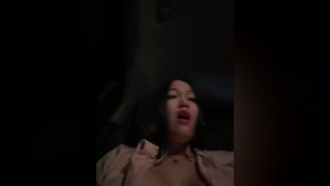 Media: A dimly-lit video of a young Asian woman with dark hair, wearing a beige robe, lying on a bed, mouth slightly open, eyes closed, expression of pleasure.