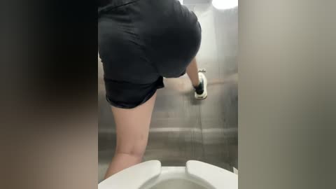 Media: A video shows a person in black shorts and a black top, squatting to flush a public restroom toilet, with a gray tiled wall in the background.