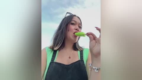 Media: Video of an Asian woman with shoulder-length dark hair, wearing a neon green tank top and black apron, licking a green popsicle, against a blurred, cloudy sky background.