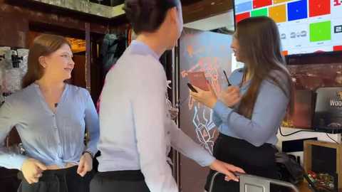 Media: Video of three women in a modern, dimly lit room. One in a blue blouse and black skirt, another in a blue blouse and black pants, and the third in a blue top and black pants, all interacting with a large screen displaying colorful graphics.