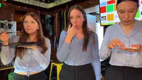 Media: Video of three young women in light blue shirts and black pants, holding phones, standing in a cluttered room with shelves, a TV, and a green chair.