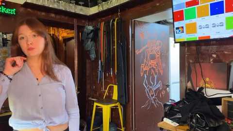 Media: Video of a woman with light skin and long brown hair in a light blue blouse, standing in a cluttered, dimly lit room with a TV, yellow stool, and colorful wall art.