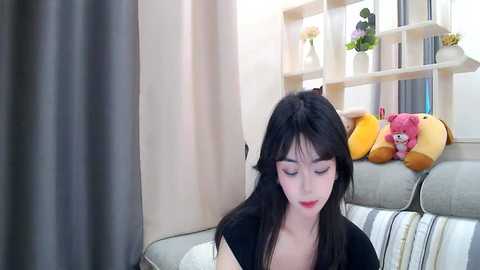 Media: Video of a young Asian woman with long black hair, wearing a black top, sitting on a light-colored sofa. Background features a window with a decorative shelf displaying stuffed toys and flowers.