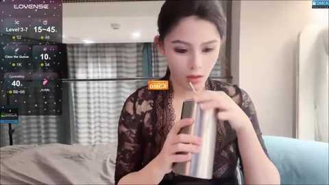 A video of a young East Asian woman with fair skin, dark hair, and wearing a black lace top, drinking from a metal straw, in a modern room with a digital display showing weather information.