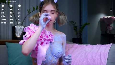 Media: Video of a young girl with blonde pigtails, wearing a light blue, frilly, ruffled dress and white gloves, applying face paint, in a modern, dimly-lit living room with plants and a sofa.