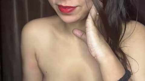 Media: Video of a young woman with fair skin, long dark hair, and red lipstick, topless, touching her neck, against a dark background.