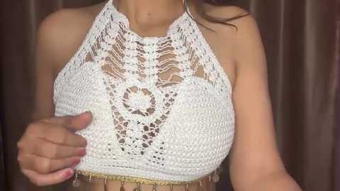 Media: Video of a woman with a light tan complexion, wearing a white, crocheted halter top with intricate floral patterns. She has long, dark hair and is indoors with brown curtains in the background.