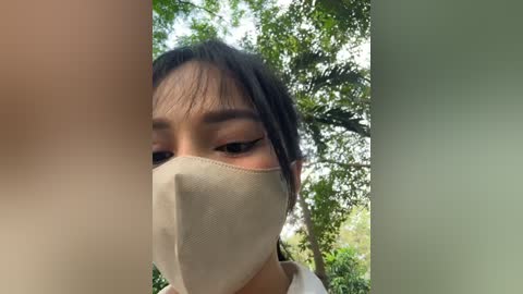 Media: A close-up video of a young Asian woman with long black hair, wearing a beige surgical mask, peering out from behind a blurred, light-colored wall. Background features green foliage and trees.