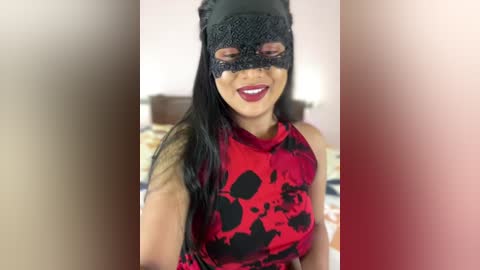 Media: Video of a smiling, dark-haired woman wearing a black lace mask and red dress with black heart pattern, set against a blurred indoor background.