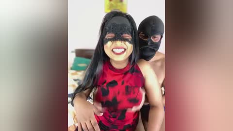 Media: Video of a smiling woman with medium skin tone and long black hair, wearing a red and black patterned top and a black mask. A shirtless man with a black mask stands behind her.