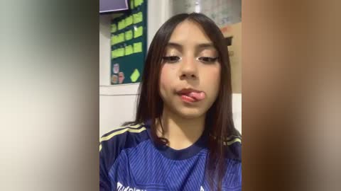 Media: Video of a young Latina woman with long dark hair, wearing a blue and yellow athletic jersey, sticking out her tongue, set in a classroom with green bulletin boards and yellow notes.