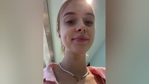 Media: Video of a young Caucasian woman with light skin and blonde hair, wearing a pink shirt and a pearl necklace, smiling, taken indoors with a soft, natural light.