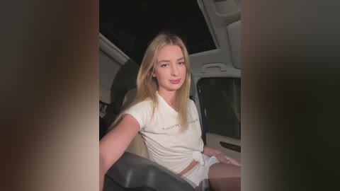 A video of a blonde woman with fair skin, sitting in a car at night, wearing a white T-shirt and shorts, looking relaxed.