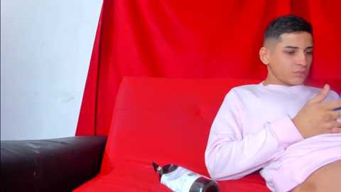 Media: Video of a young Latino man with short dark hair in a light pink shirt, sitting on a red couch, holding a bottle of alcohol. Background is a red curtain.