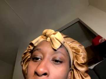 Media: Video of a Black woman with medium brown skin and a headscarf tied in a bow, wearing minimal makeup, in a dimly lit room with a mirror and a partially visible red bag in the background.