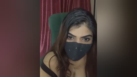 Media: Video of a young woman with long, dark hair, wearing a black face mask and a black off-shoulder top, seated in a dimly lit room with maroon and dark brown walls.