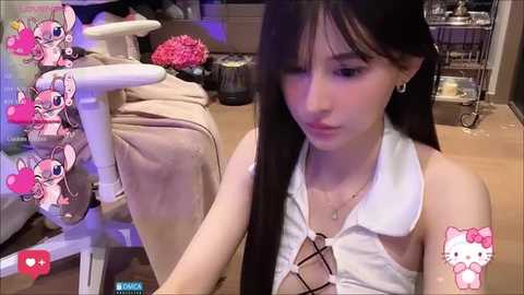 Media: Video of an East Asian woman with long black hair in a white sleeveless top, seated in a cluttered room with a white chair and pink stuffed animal.