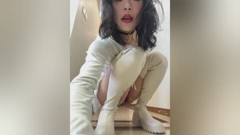 Media: Video of a woman with pale skin, black hair, and red lipstick, wearing a white long-sleeve top, white thigh-high stockings, and white boots, squatting on a wooden floor with a white wall in the background.