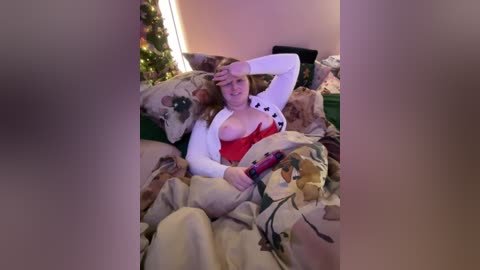 Media: Video of a woman in a red top, white cardigan, and beige blanket, with a Christmas tree in the background, lying on a couch.