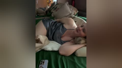 Media: Video of a white woman with short blonde hair, wearing a grey tank top, lying on a green bedspread in a dimly lit bedroom.