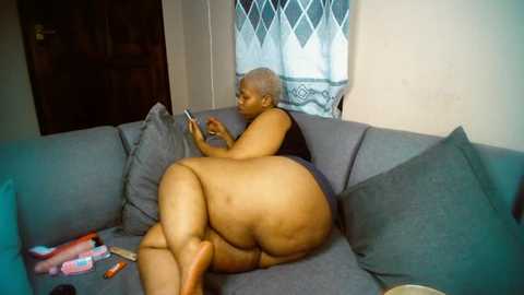 Media: Video of an elderly, obese Black woman with short gray hair, nude, lying on a gray sofa, playing with a smartphone, surrounded by pillows and scattered items.