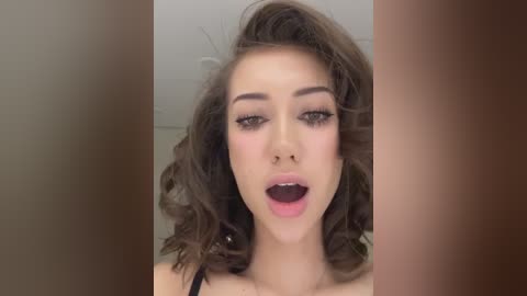 Media: Video of a young woman with light skin and voluminous, wavy brown hair. She has dark, dramatic eyeliner and full lips slightly parted, giving a sensual expression. The background is blurred, with vertical brown and beige tones.