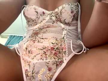 Media: Video of a woman in a sheer, pastel pink lingerie bodysuit adorned with floral embroidery, sitting with legs spread, revealing her toned thighs and smooth skin. Background shows a blurred, indoor setting with a blue curtain.