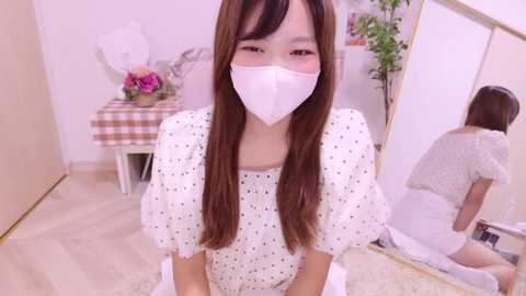 Media: Video of an East Asian woman with long brown hair, wearing a white mask and polka-dot dress, kneeling in a bright, minimalistic room with a mirror and plants.