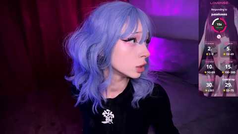 Video of a pale-skinned, slender woman with shoulder-length, wavy, light blue hair, wearing a black top, in a dimly lit room with purple lighting. A screen overlay shows her as an anime character.