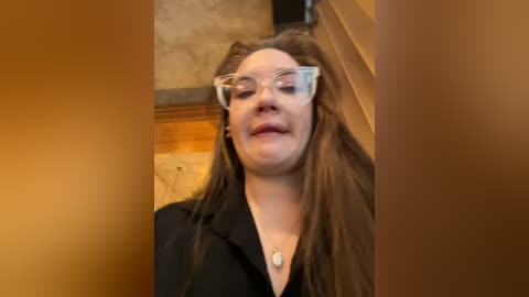 Media: Video of a smiling woman with long brown hair, wearing large glasses and a black coat, standing indoors with a wooden staircase in the background.