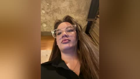 Media: Video of a light-skinned woman with long brown hair, wearing clear, oversized eyeglasses and a black shirt, standing in a dimly lit room with a textured gray wall and wooden paneling.