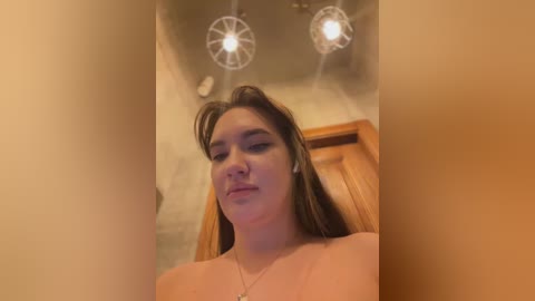 Media: Video of a young woman with fair skin, brown hair, and a small nose piercing, sitting in a wooden chair with two ceiling lights.