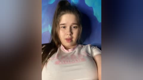 Video of a young, fair-skinned girl with long brown hair, wearing a white t-shirt with pink text, against a blue and purple backdrop.