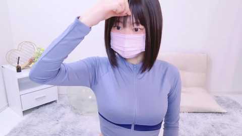 Media: Video of an East Asian woman with shoulder-length black hair, wearing a light blue zip-up top and a pink face mask, adjusting her hair in a bright, minimalist room with white walls and a white couch.