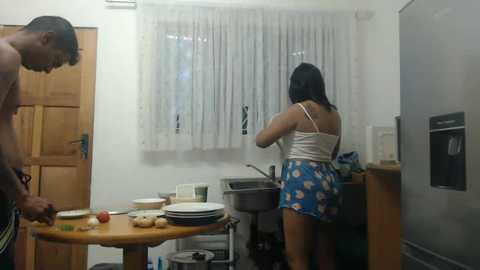 Media: Video of a kitchen with a woman in a white tank top and blue floral shorts washing dishes. A man in shorts stands beside a wooden table.